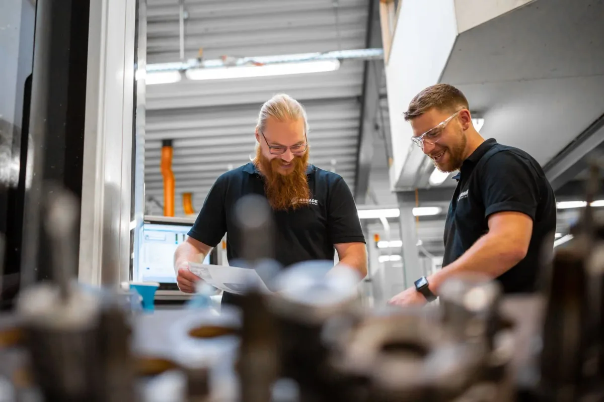 Zwei Mitarbeiter der Schade GmbH prüfen lachend eine Zeichnung in der Fertigungshalle